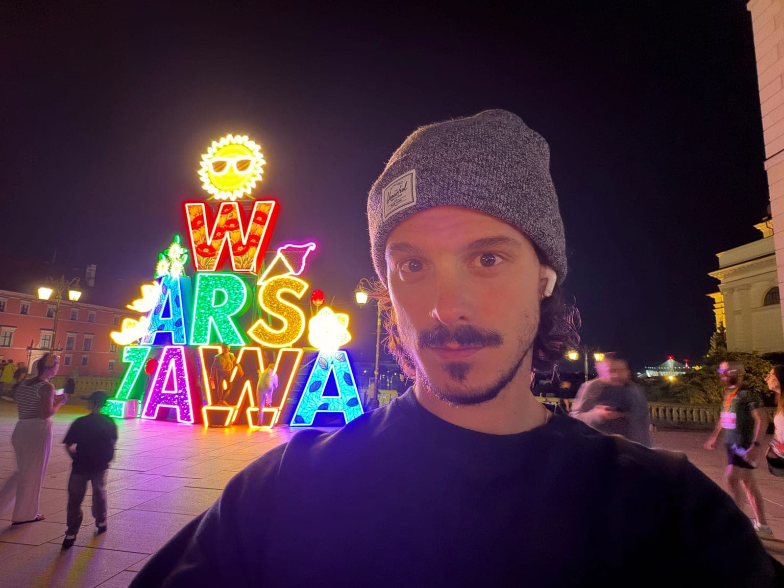 Joshua at night in Warsaw's Old Town in front of a neon WARSZAWA sign